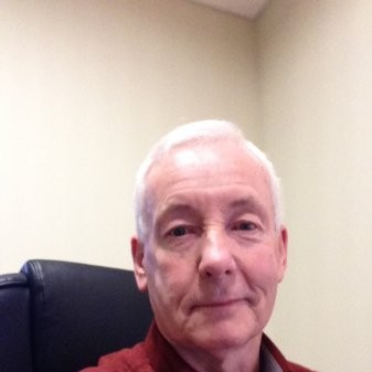Portrait of an older man with short white hair sitting in a black chair against a beige wall.