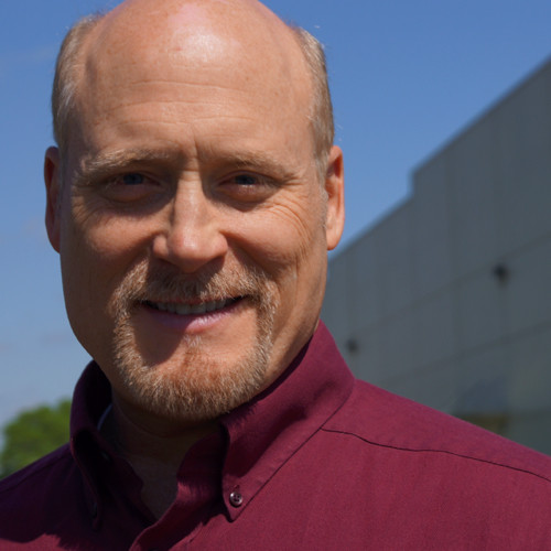 Smiling man with a bald head and goatee wearing a maroon button-up shirt outdoors.