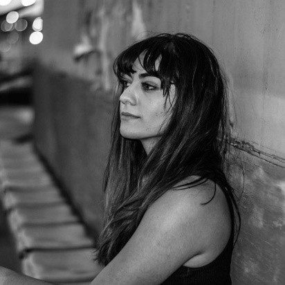 Black and white portrait of a woman with long dark hair and bangs sitting sideways against a wall.