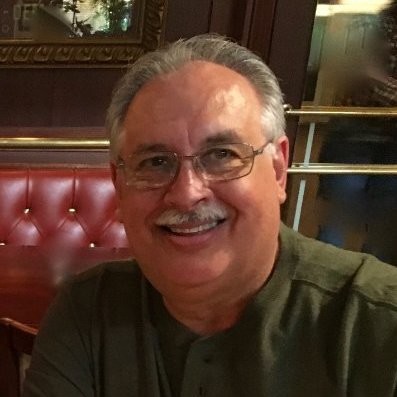 Smiling middle-aged man with gray hair, glasses, and a mustache sitting indoors with a red cushioned bench behind him.