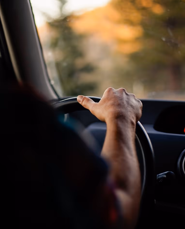 Close-up of a person's hand gripping a car steering wheel while driving at sunset.