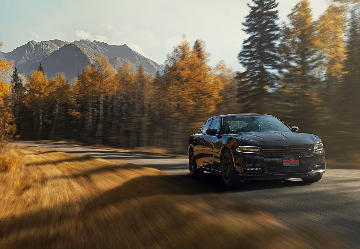 Black car driving on road with autumn trees and mountains in background under a clear sky.