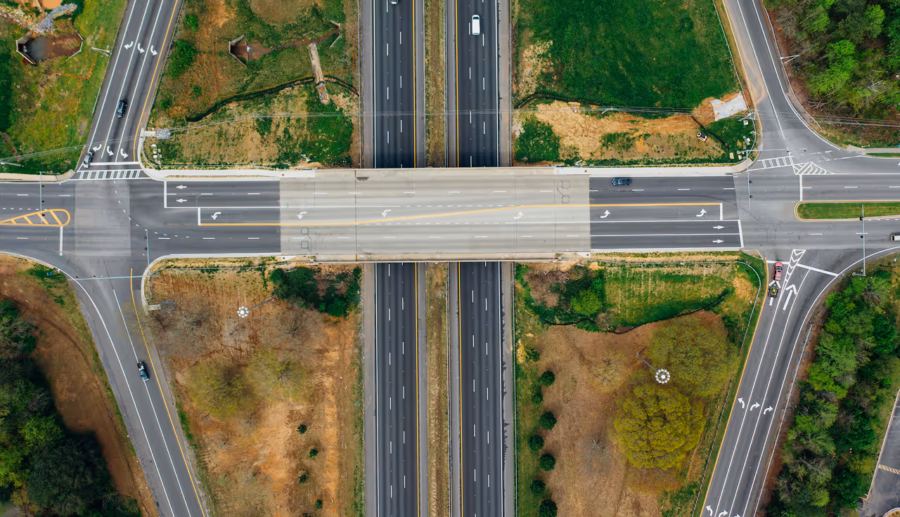 Aerial view of a highway interchange with cars, roads, green and brown grassy areas around it.