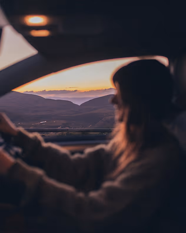 Silhouette of a woman driving a car with a mountain sunset visible through the window.