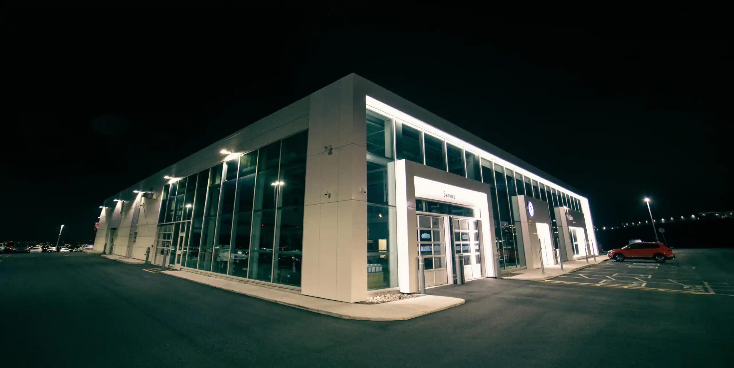 Modern car dealership building with large glass windows lit up at night with parking spaces and one red car.
