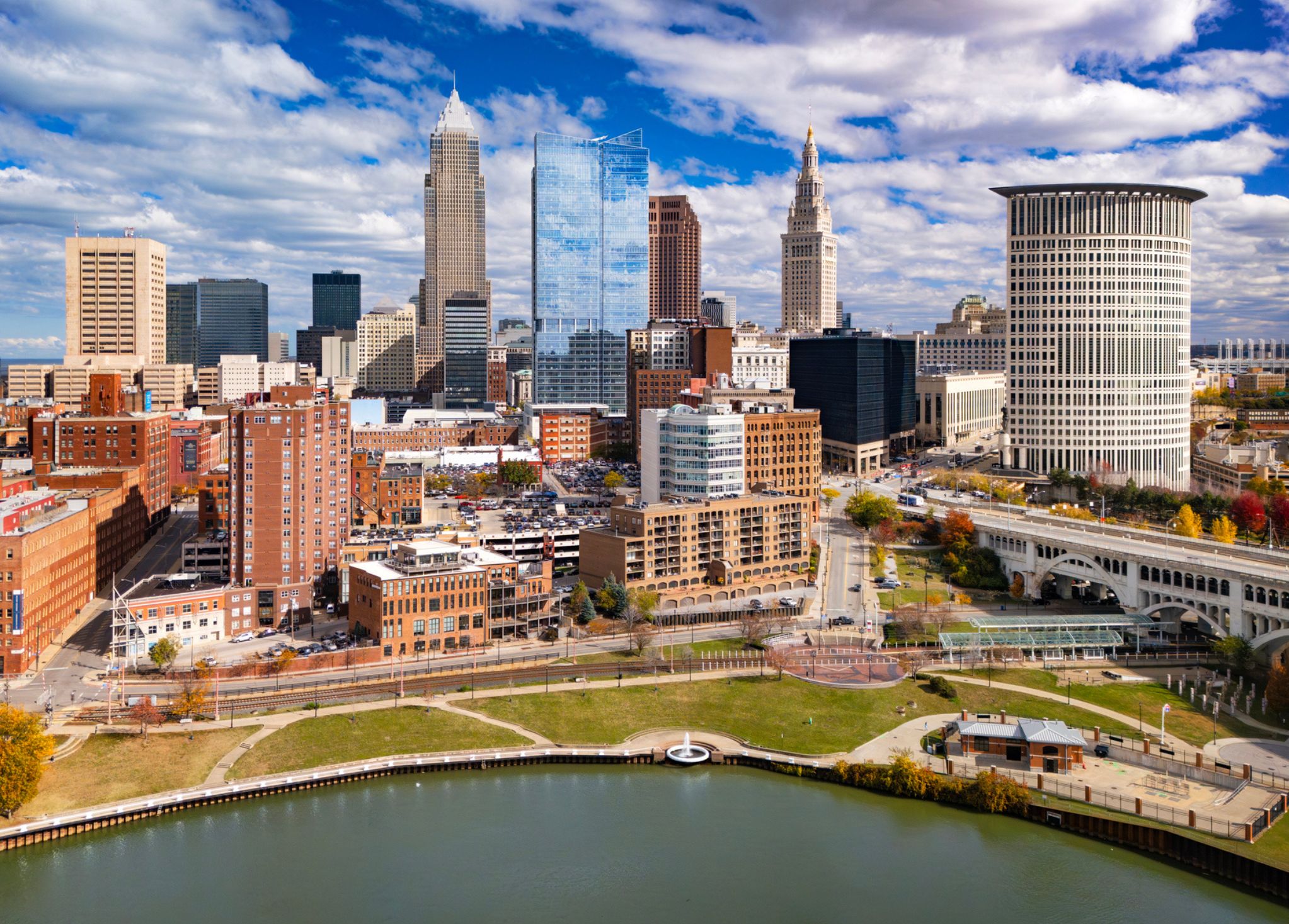 Downtown city skyline with tall buildings, a riverfront park, and partly cloudy sky.