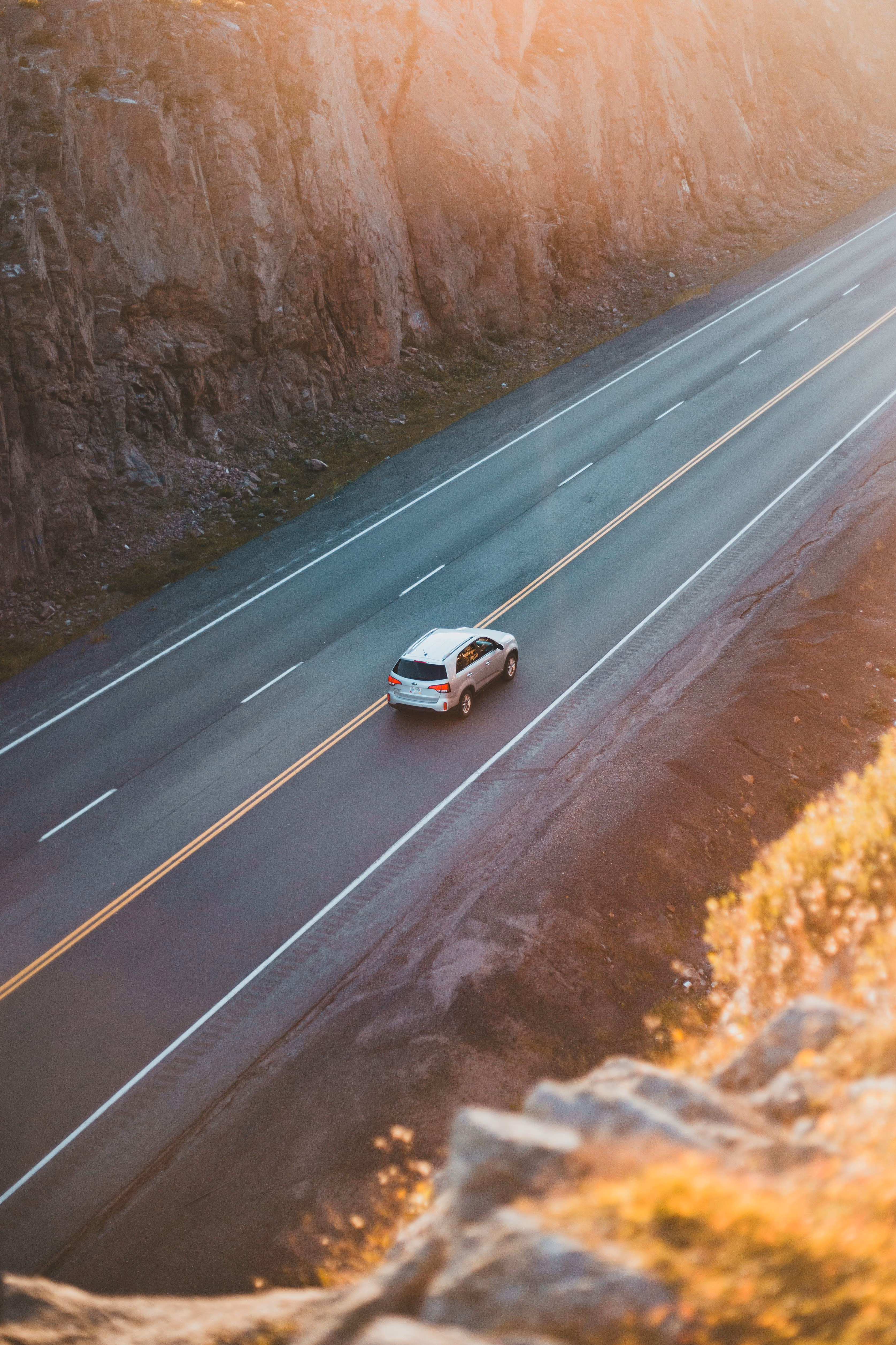 White SUV driving on a two-lane road beside rocky cliffs at sunset.