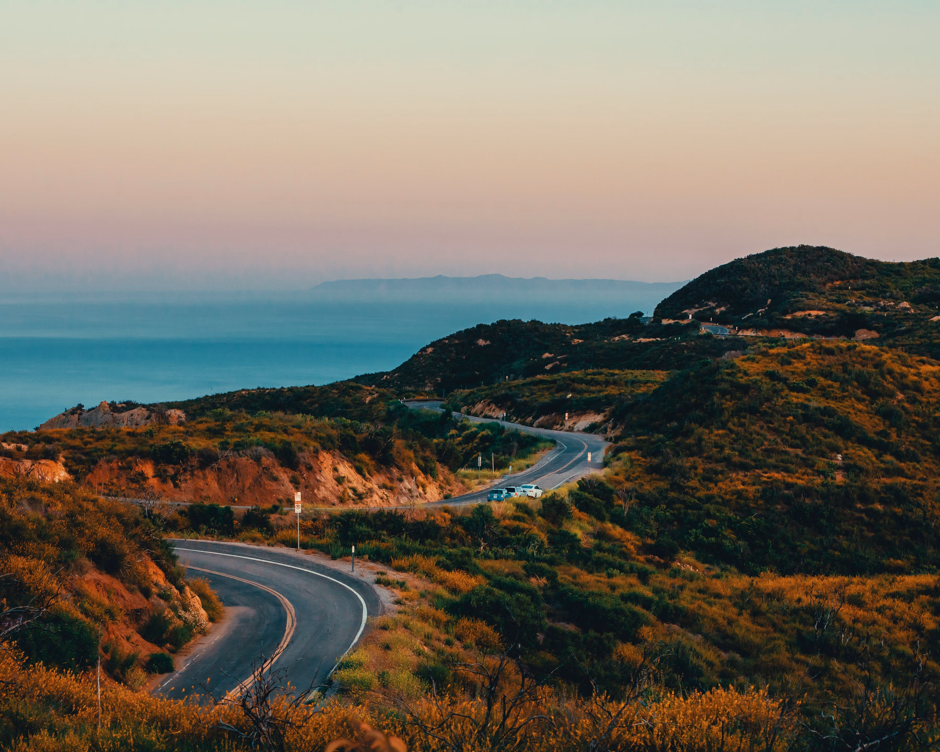 Winding road through green hills with a few parked cars under a pastel sky near the coast.