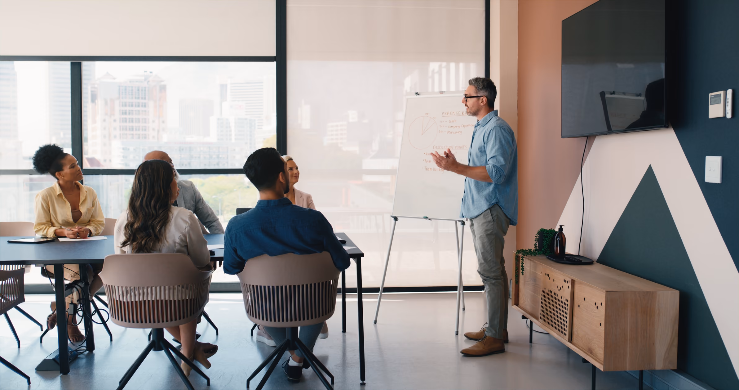 Man in glasses presenting to a group of five seated colleagues in a modern office with city view.