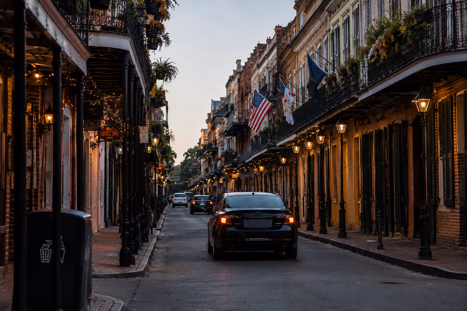 Street at dusk with old buildings, hanging plants, and cars driving under glowing street lamps.