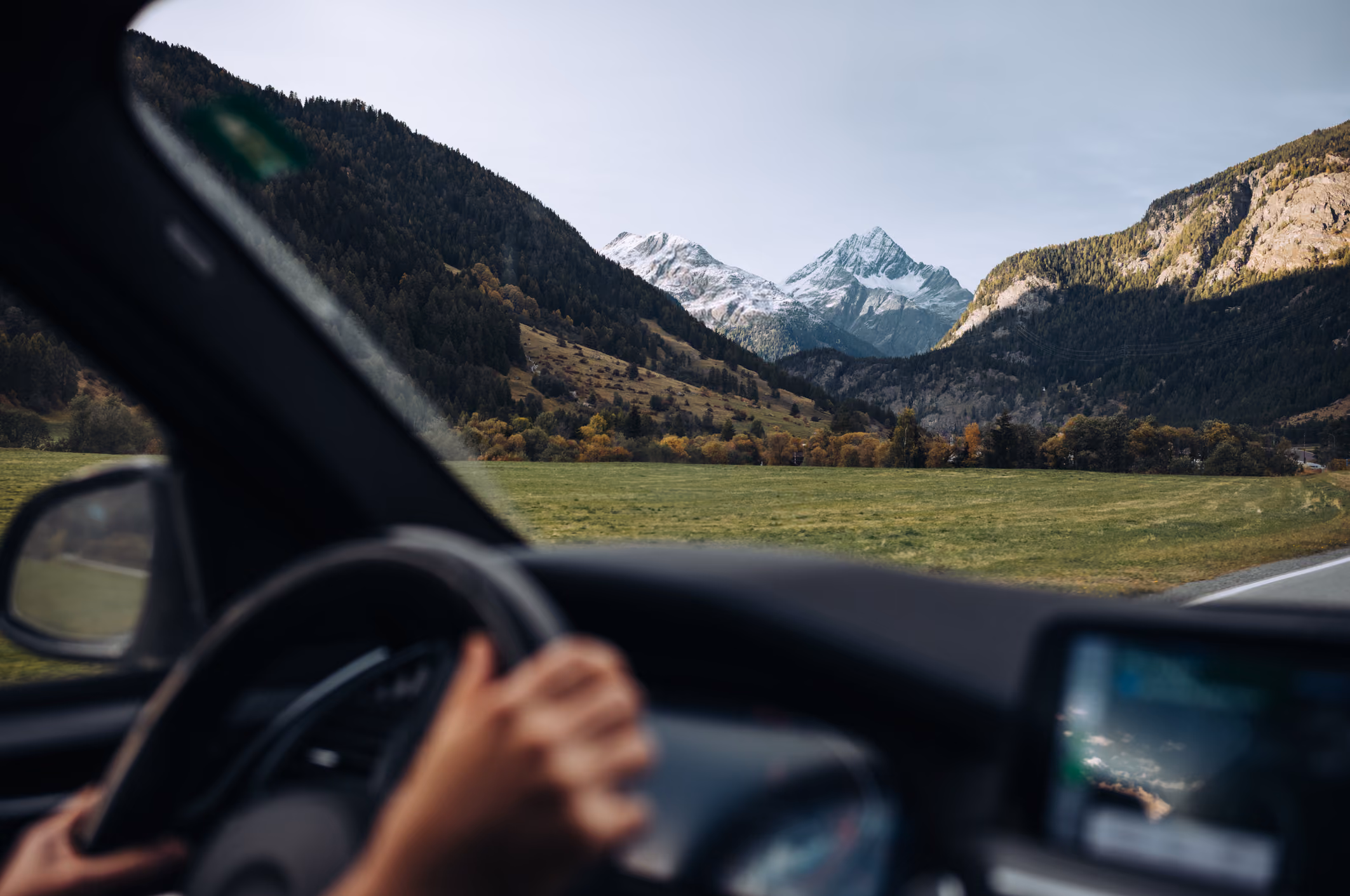 View of snow-capped mountains and green valley from inside a car with hands on the steering wheel.