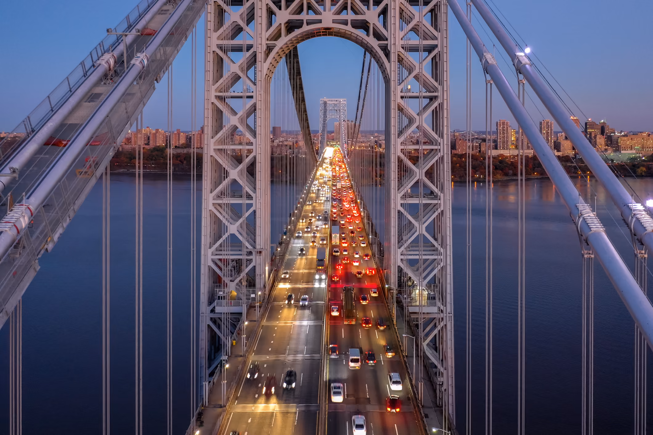 Traffic moving on a large suspension bridge over a river with city skyline at dusk.
