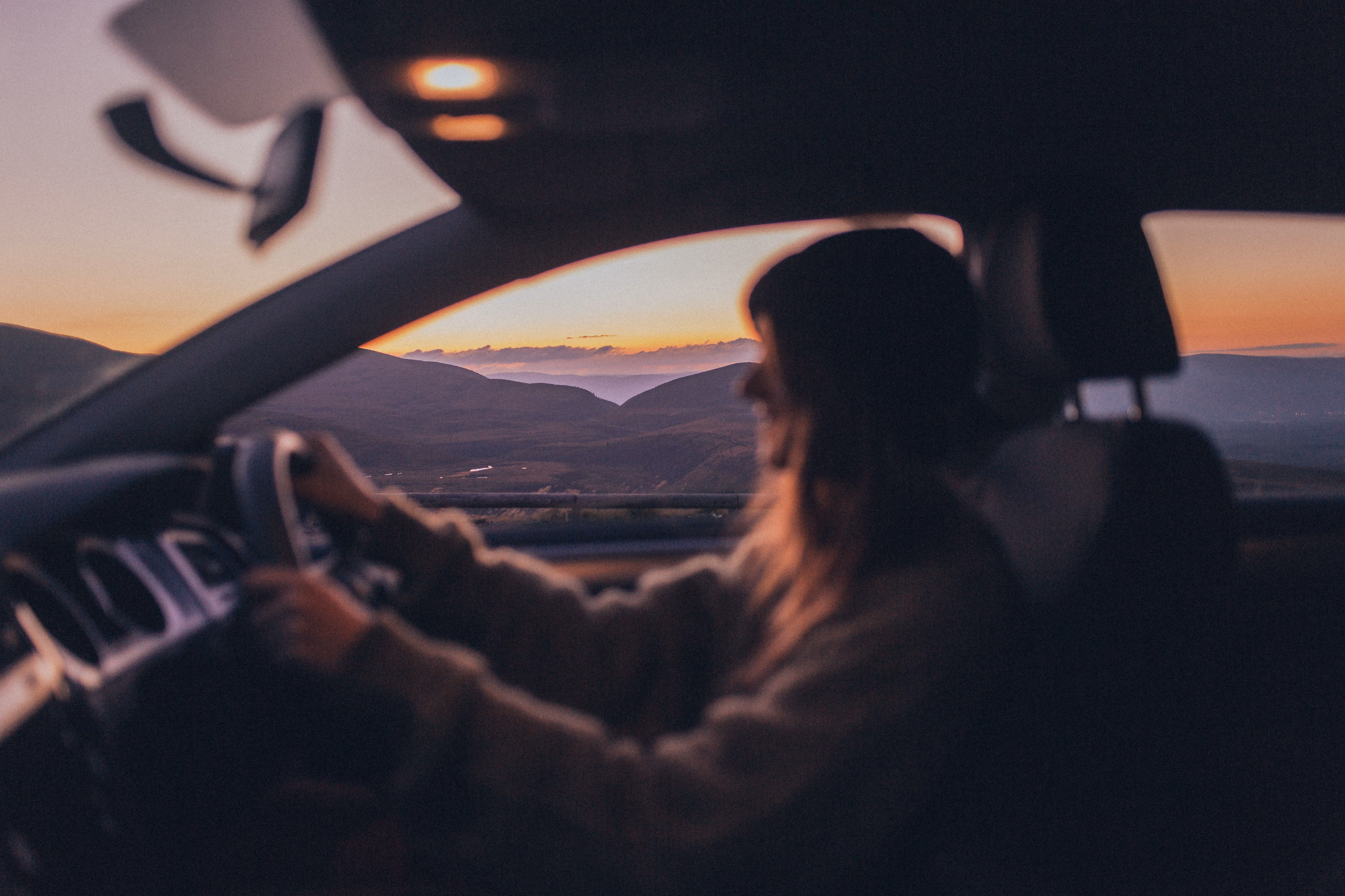 Silhouette of a person driving a car with a mountain landscape at sunset in the background.