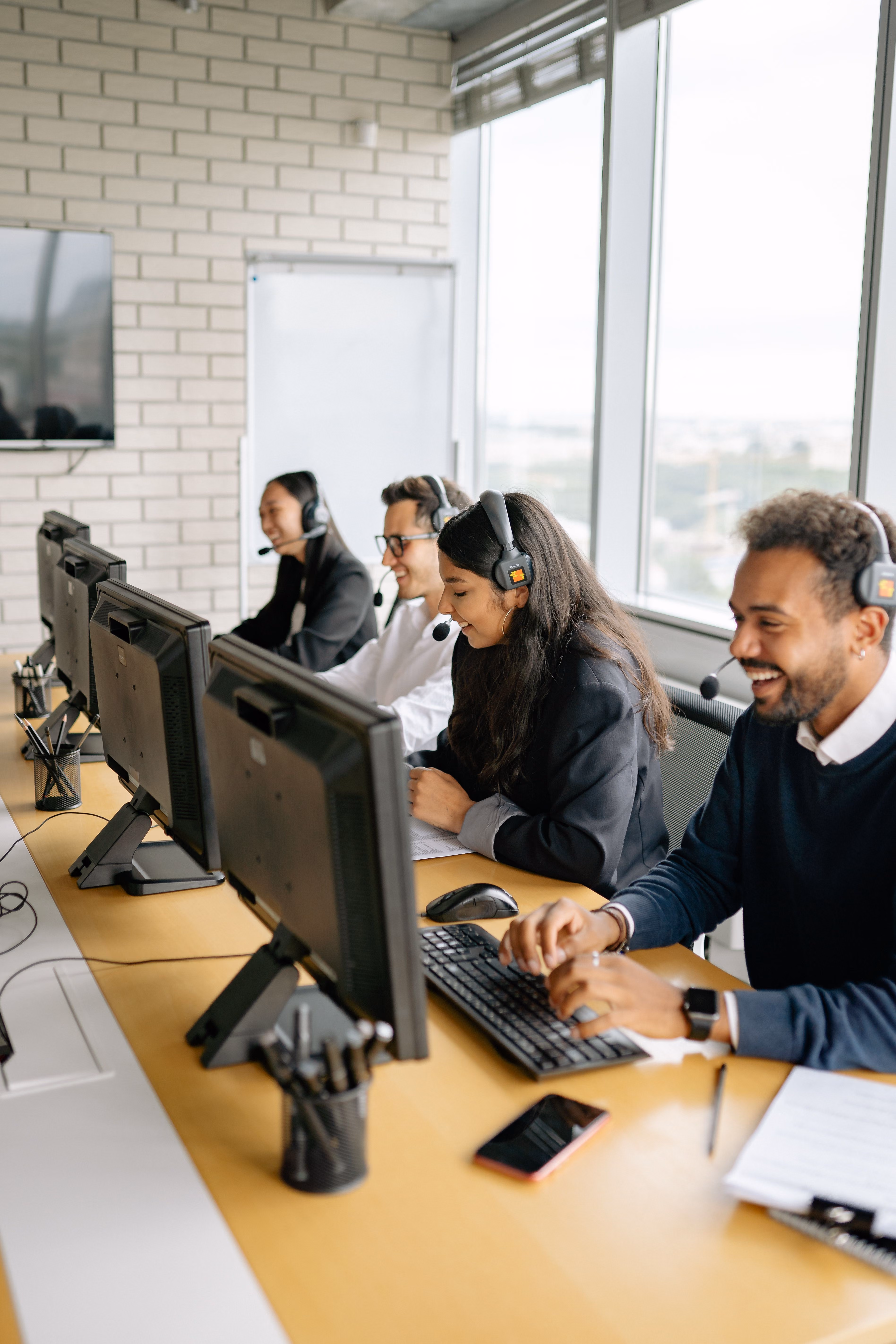 Four call center agents wearing headsets working at computers in a bright office with large windows.