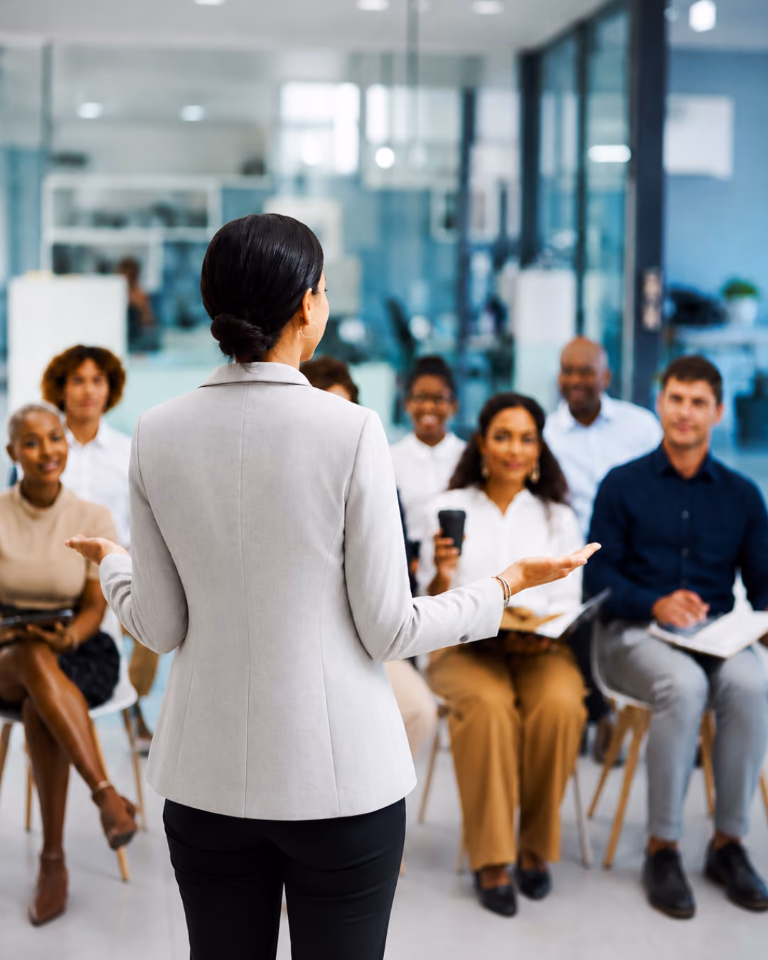 Businesswoman in gray blazer presenting to a diverse seated audience in a modern office.