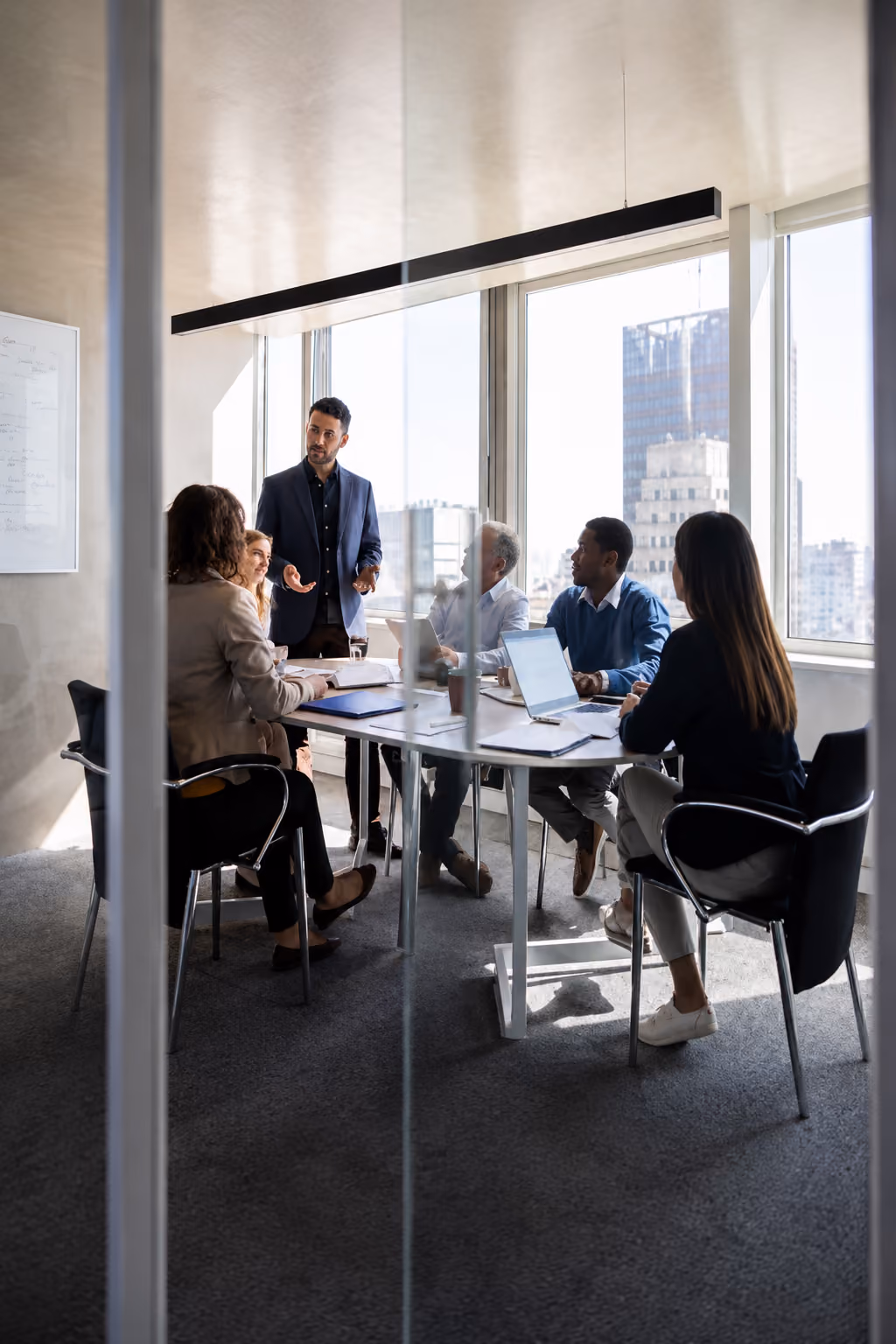 Five diverse coworkers in a bright office having a meeting around a table with laptops and papers.