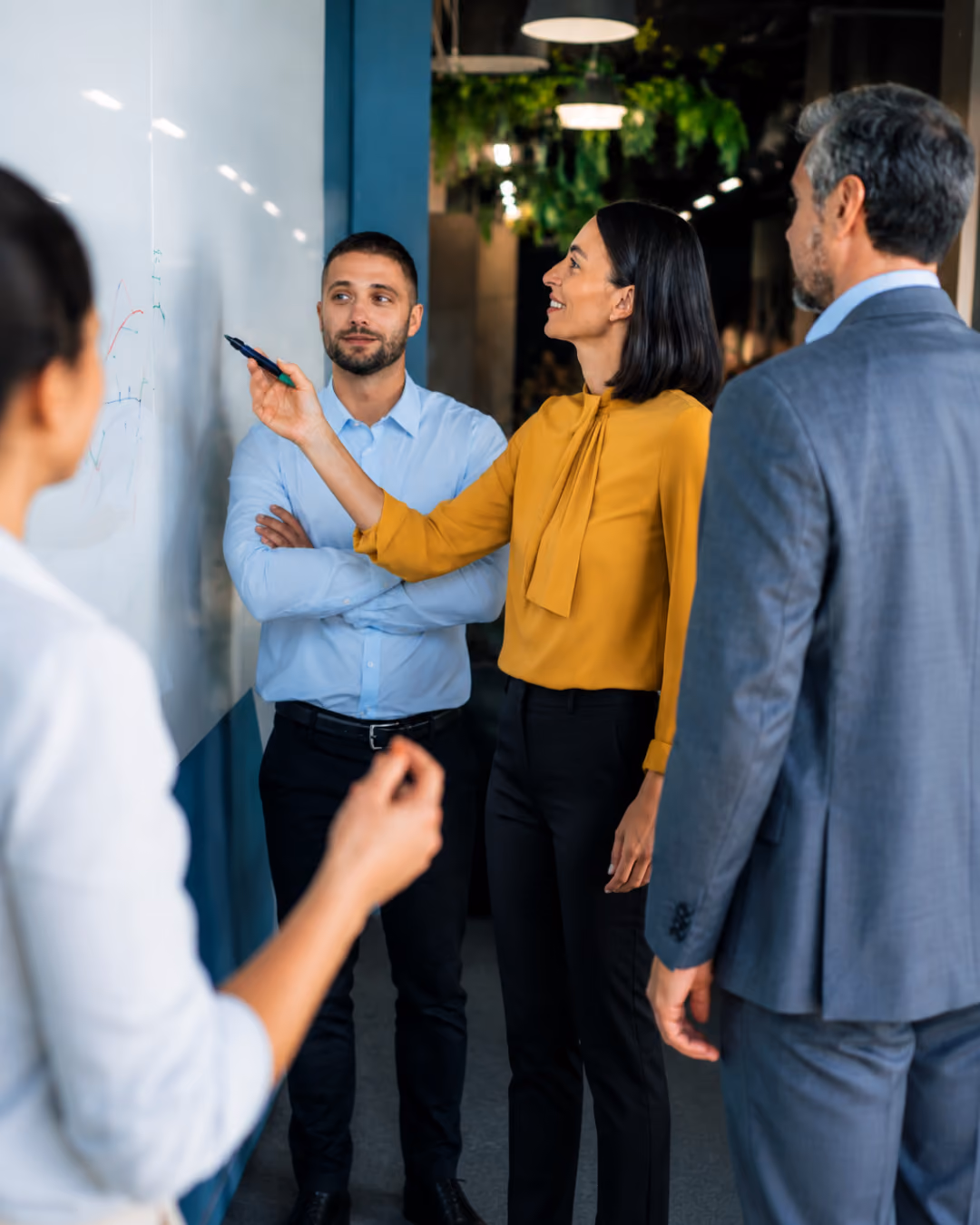 Four businesspeople discuss and point to charts on a whiteboard in a modern office.
