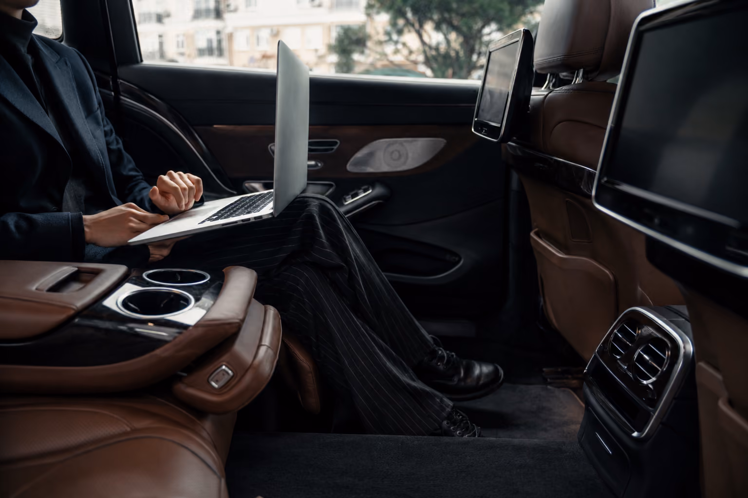 Person in business attire working on a laptop inside a luxury car's brown leather backseat.
