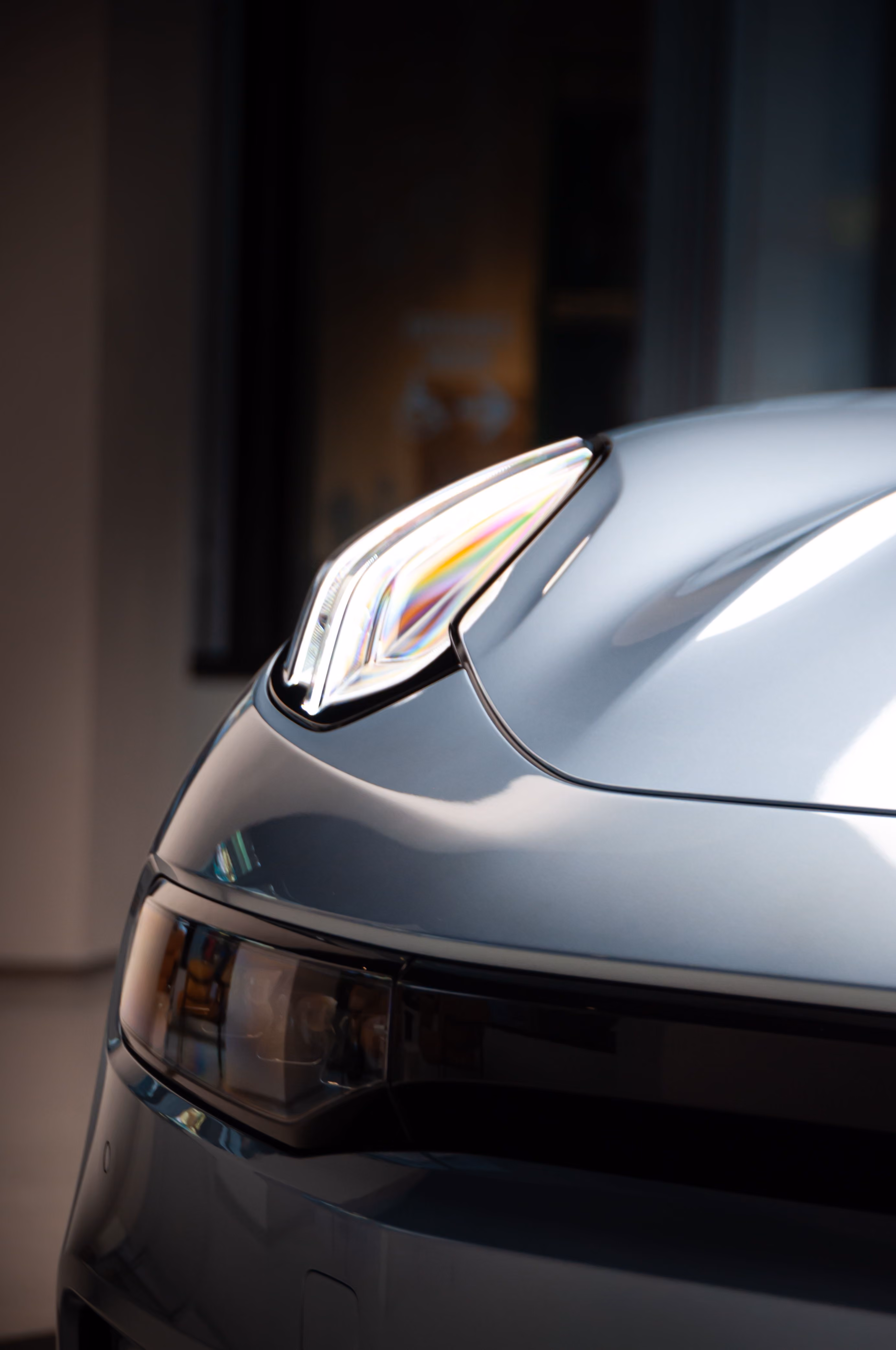 Close-up of a silver car's front headlight and sleek hood design in soft indoor lighting.