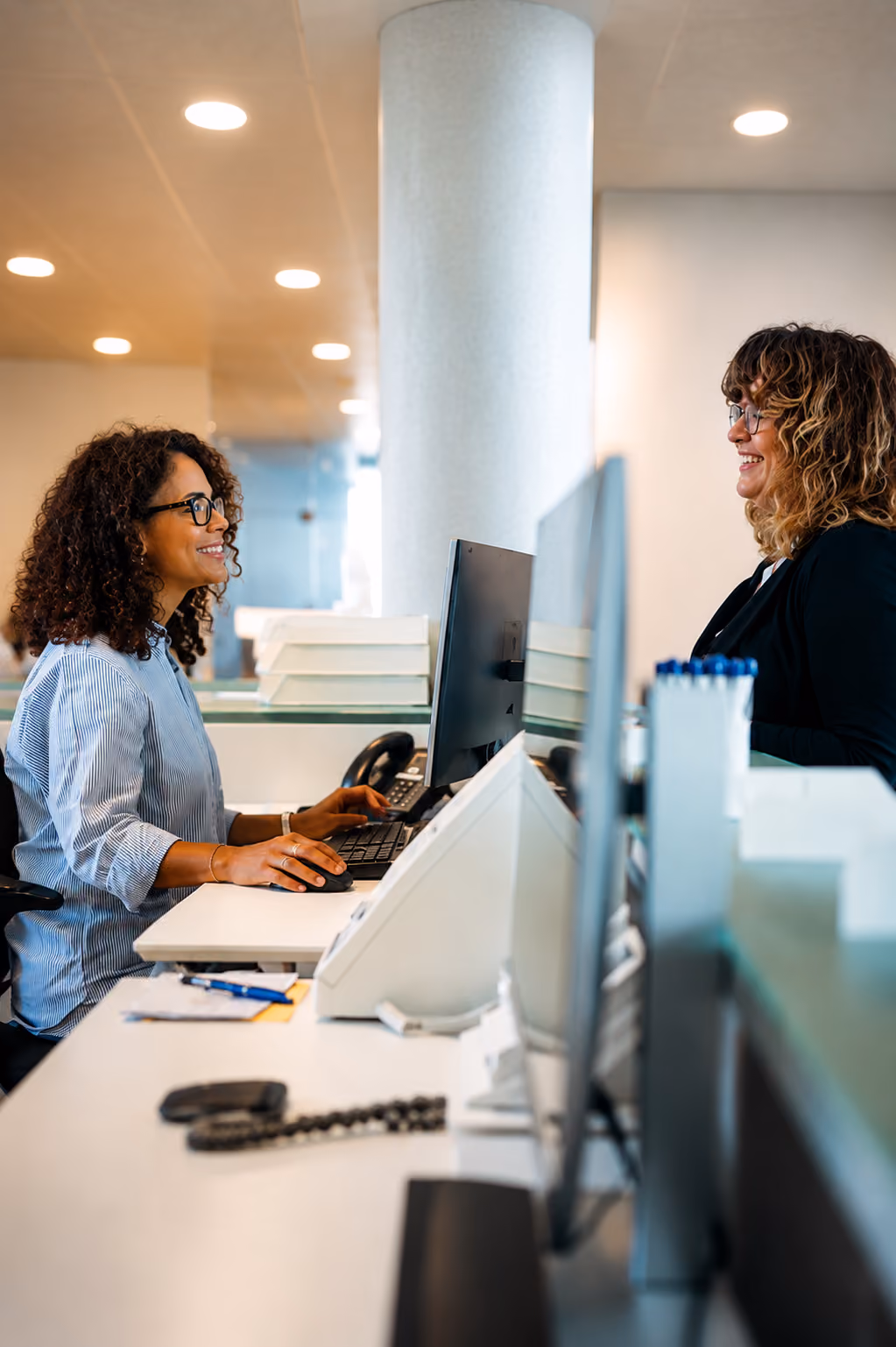 Two women smiling and talking across an office desk with computer monitors between them.
