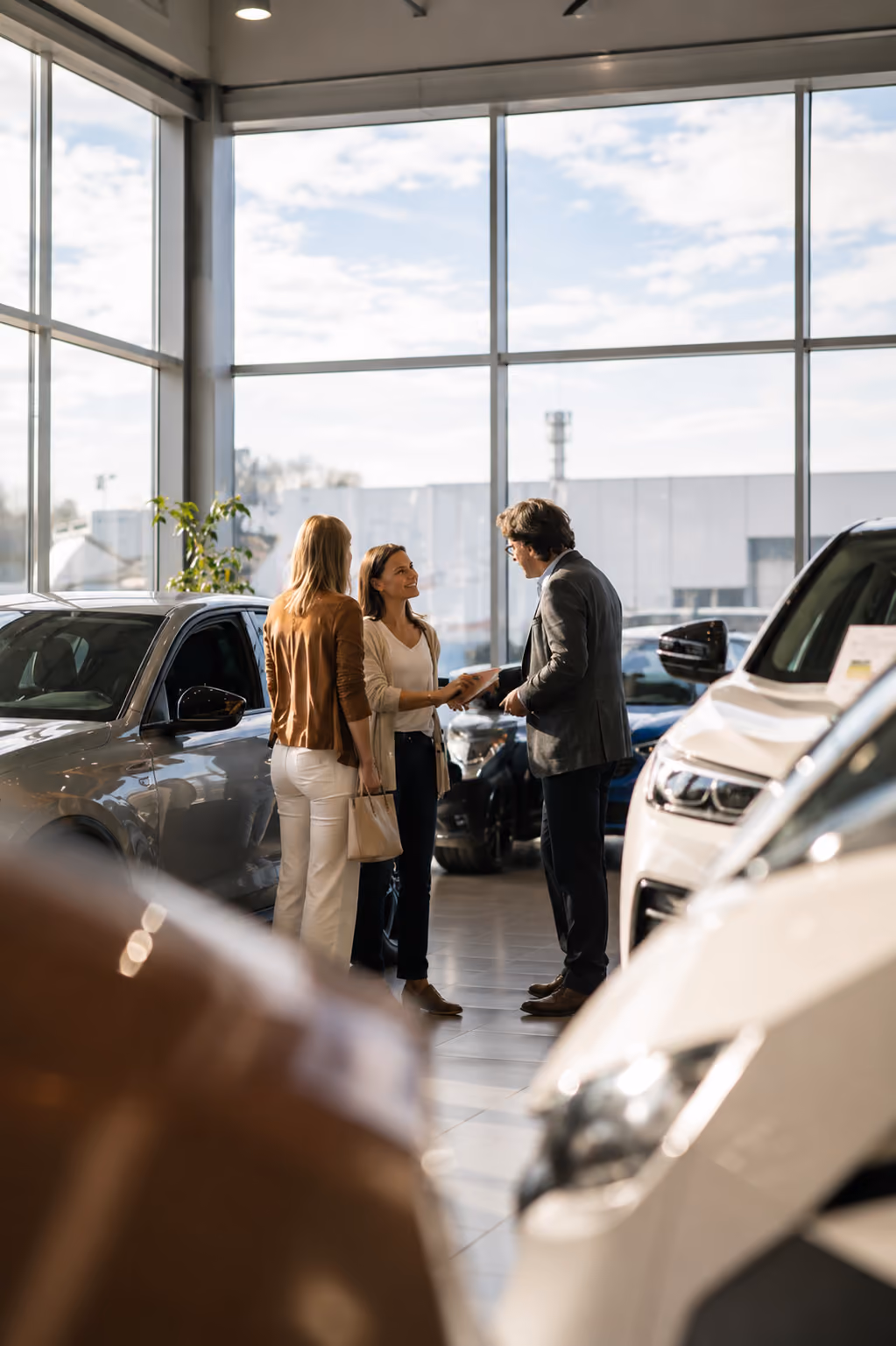 Two women talk with a man in a car dealership showroom surrounded by cars and large windows.
