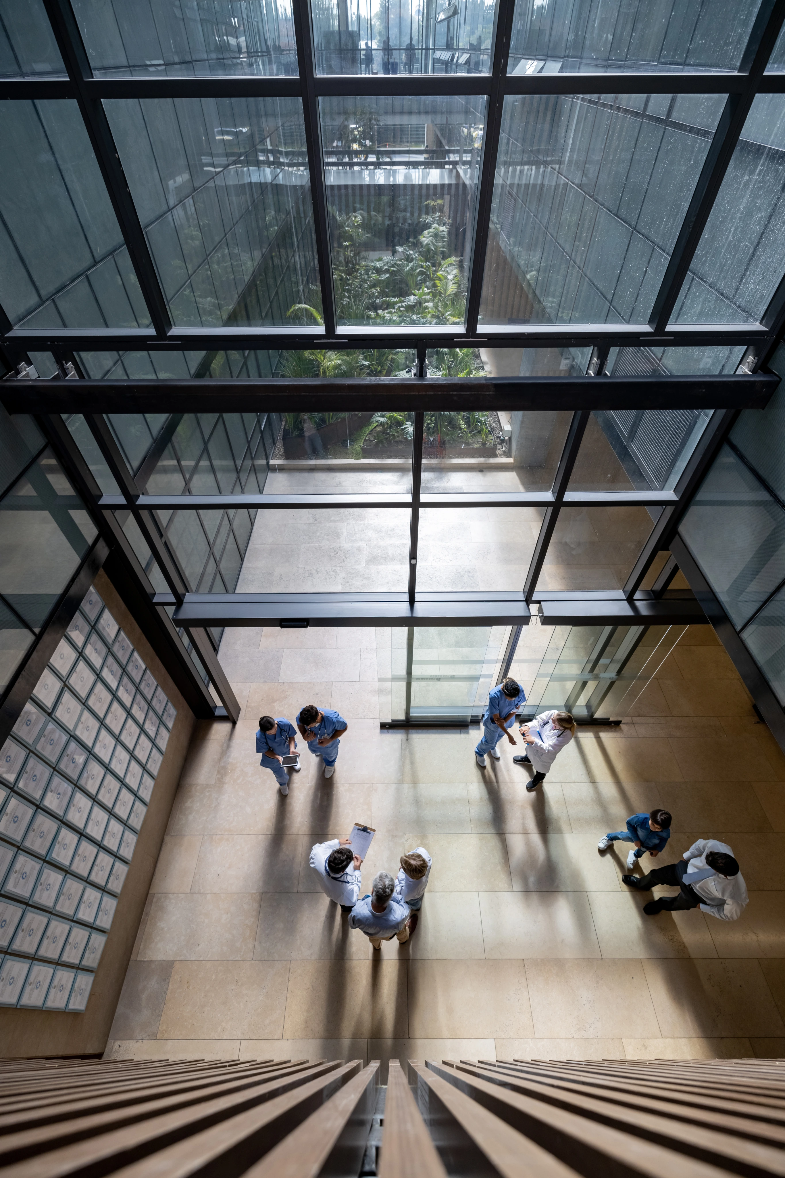 Aerial view of medical professionals and staff standing and talking in a modern hospital lobby with large glass windows and tiled floor.