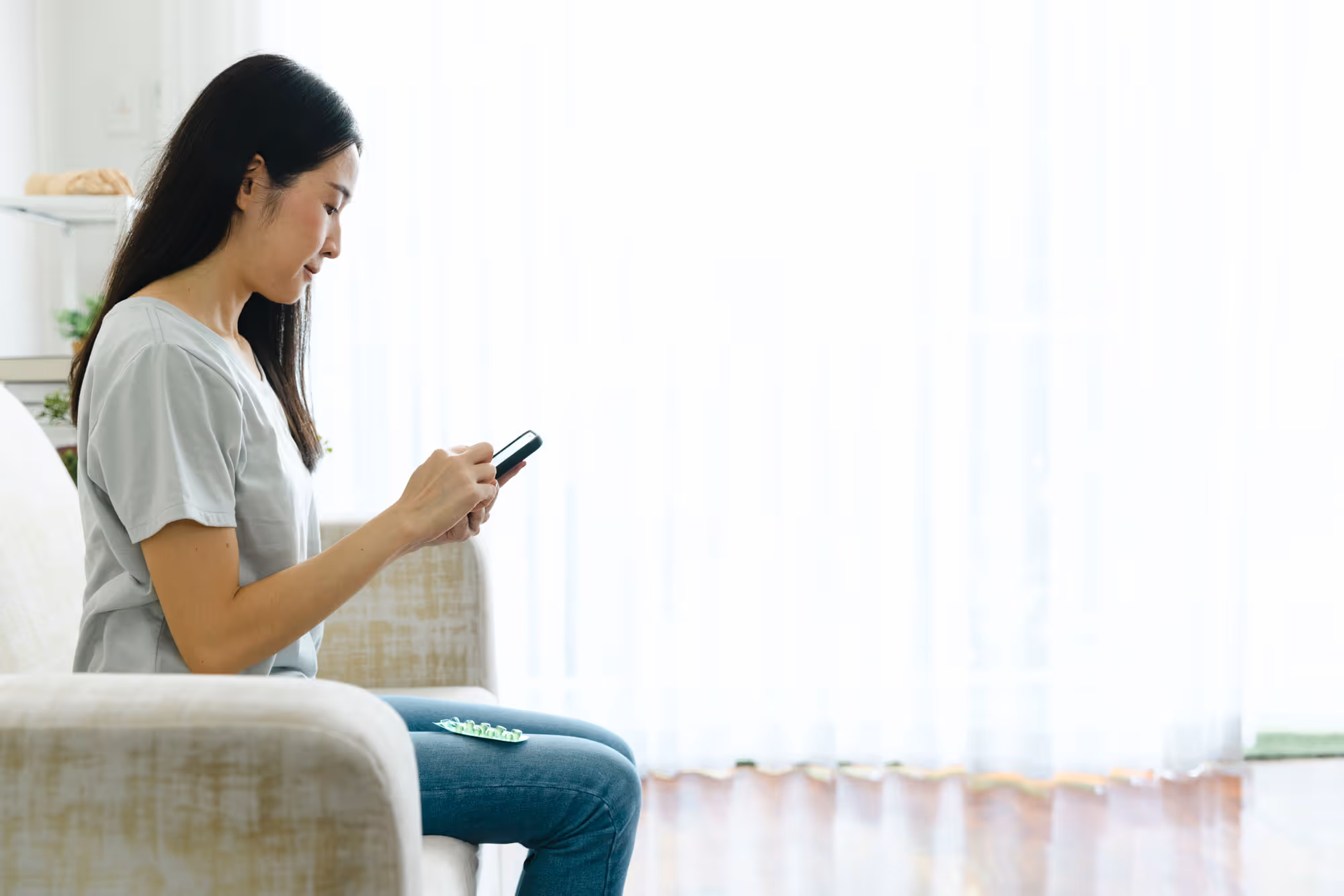 Woman sitting on a couch using a smartphone with a blister pack of pills on her lap in a bright room.