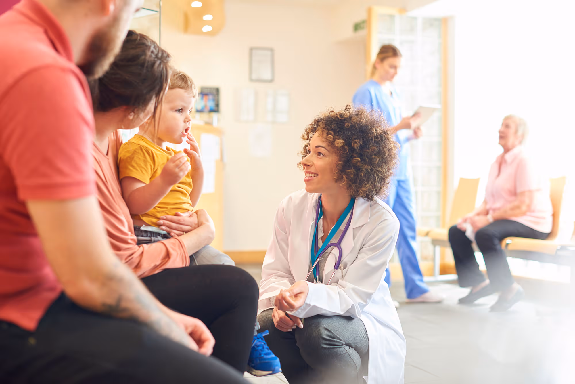 A female doctor with a stethoscope kneels and smiles while talking to a young child sitting on a woman's lap in a medical waiting room.