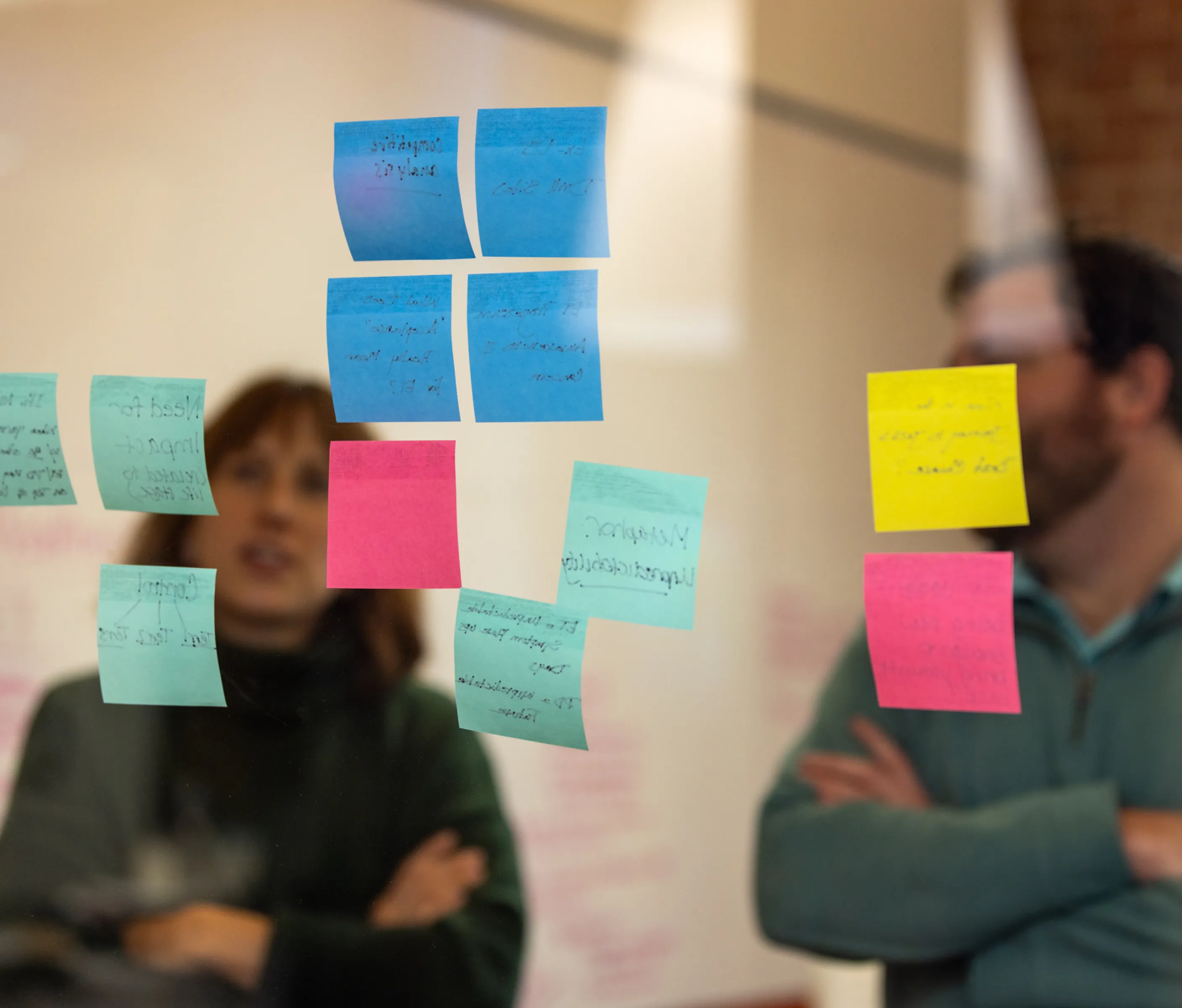 Two people standing behind a glass wall covered with colorful sticky notes.