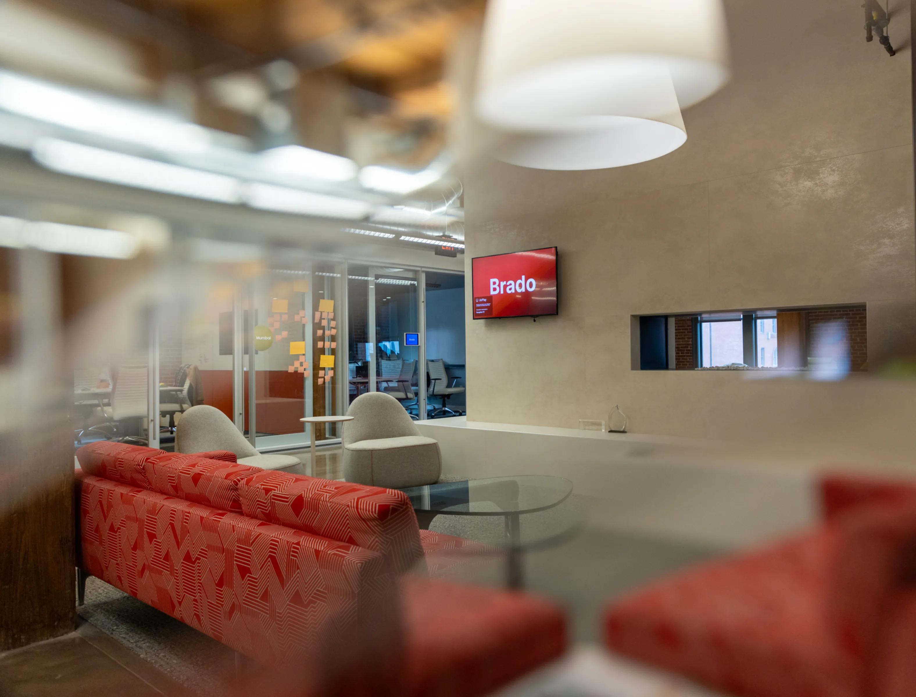 Modern office lounge with red patterned sofa, two gray chairs, glass coffee table, and a wall-mounted screen displaying 'Brado'.