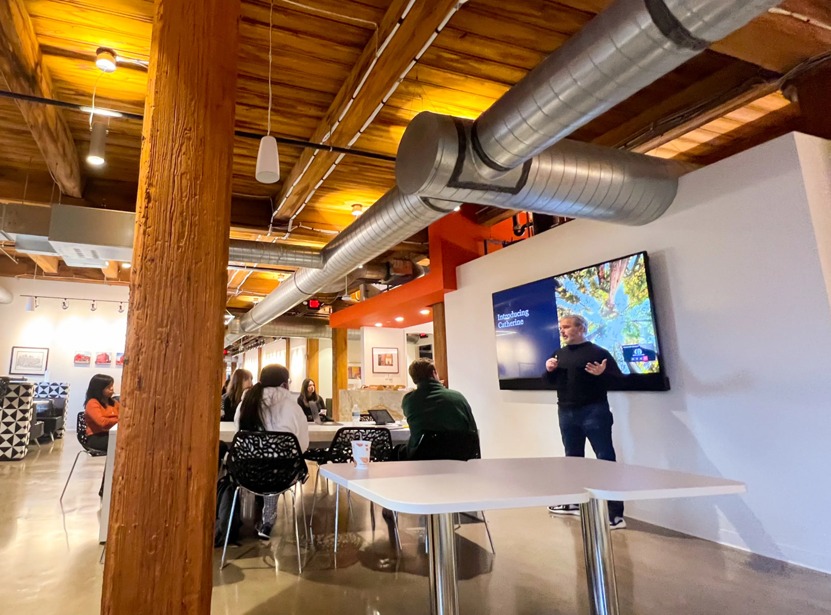 A man giving a presentation to a seated group in a modern room with wooden beams and exposed ductwork.