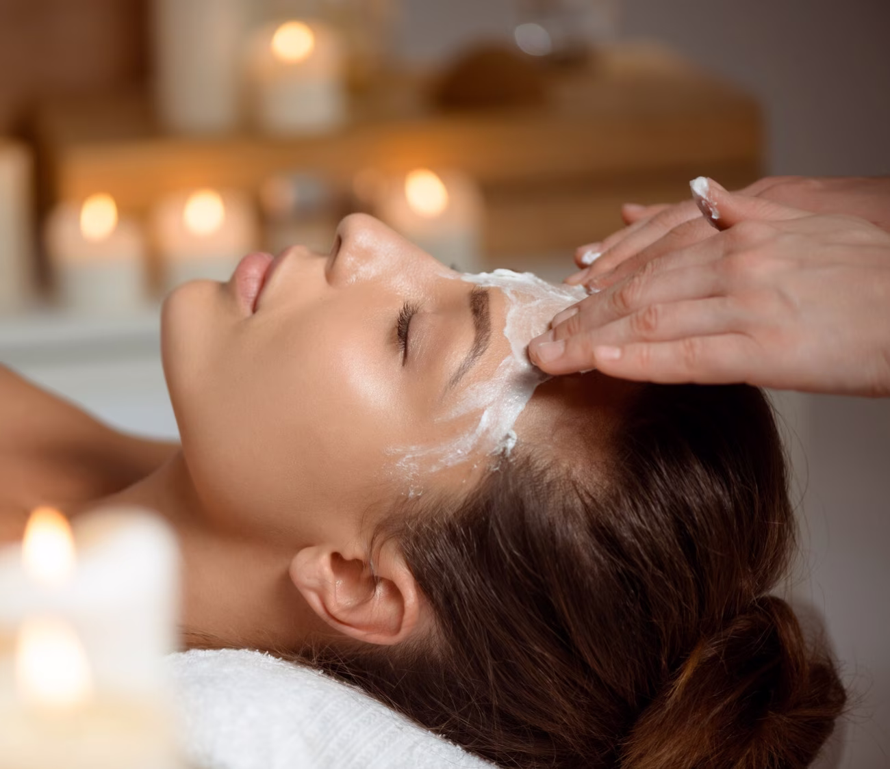 Woman receiving a facial treatment with cream applied on forehead in a spa setting with lit candles.