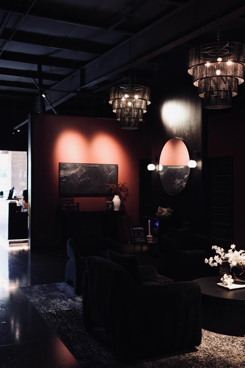 Calm massage room with black-covered massage table, beige towel, black curtains, white chair, and decorative plants.