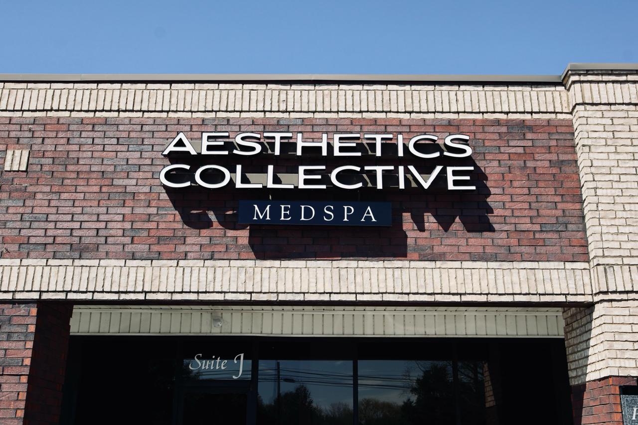 Brick building with white columns and a sign that reads 'Aesthetics Med Collective Spa' above a white door and windows, with two empty parking spaces in front.