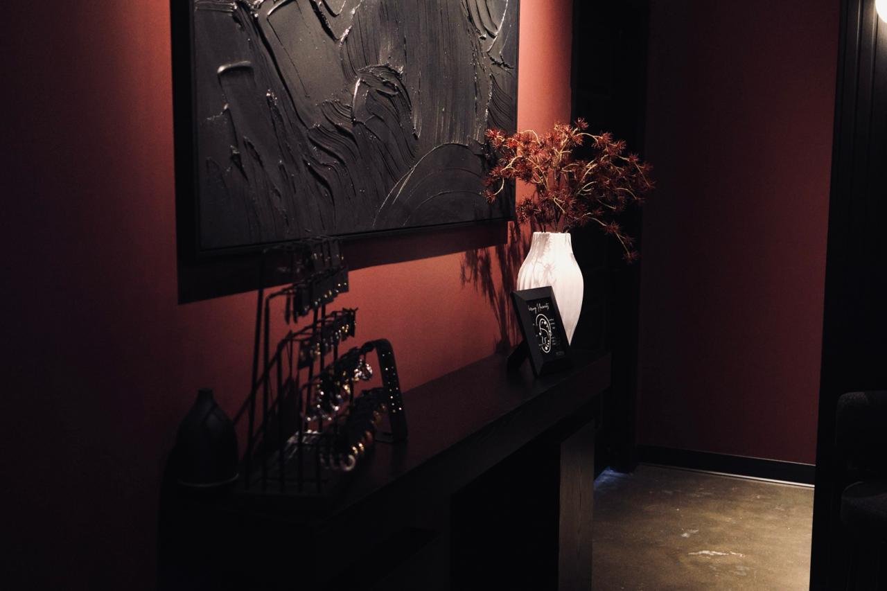 Calming treatment room with a massage table covered in dark and white towels, a black cabinet, a standing lamp, and shelves with skincare products.