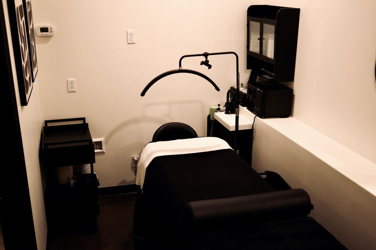 Minimalist spa room with black massage table, white chair, marble floor, and two hanging woven pendant lights.
