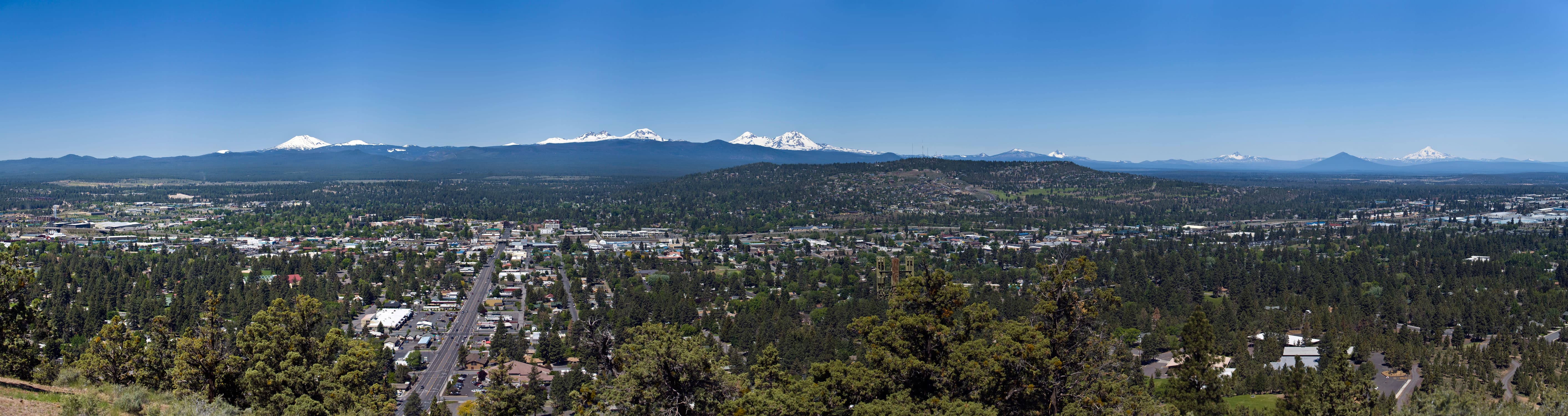 Panoramic view of a city surrounded by forests with snow-capped mountains under clear blue sky in the background.