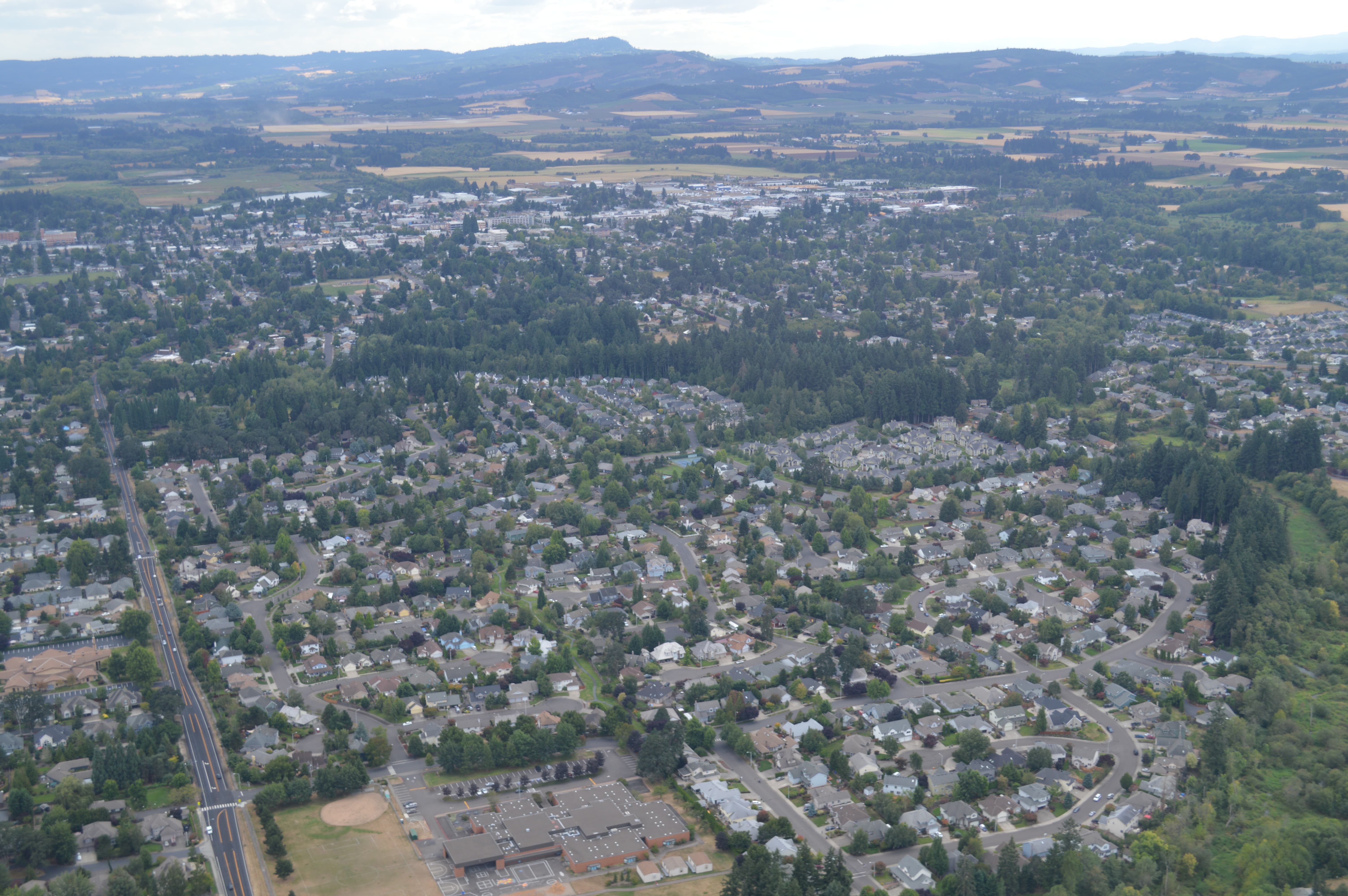 Aerial view of a suburban neighborhood with houses, trees, streets, a school building, and surrounding hills in the distance.