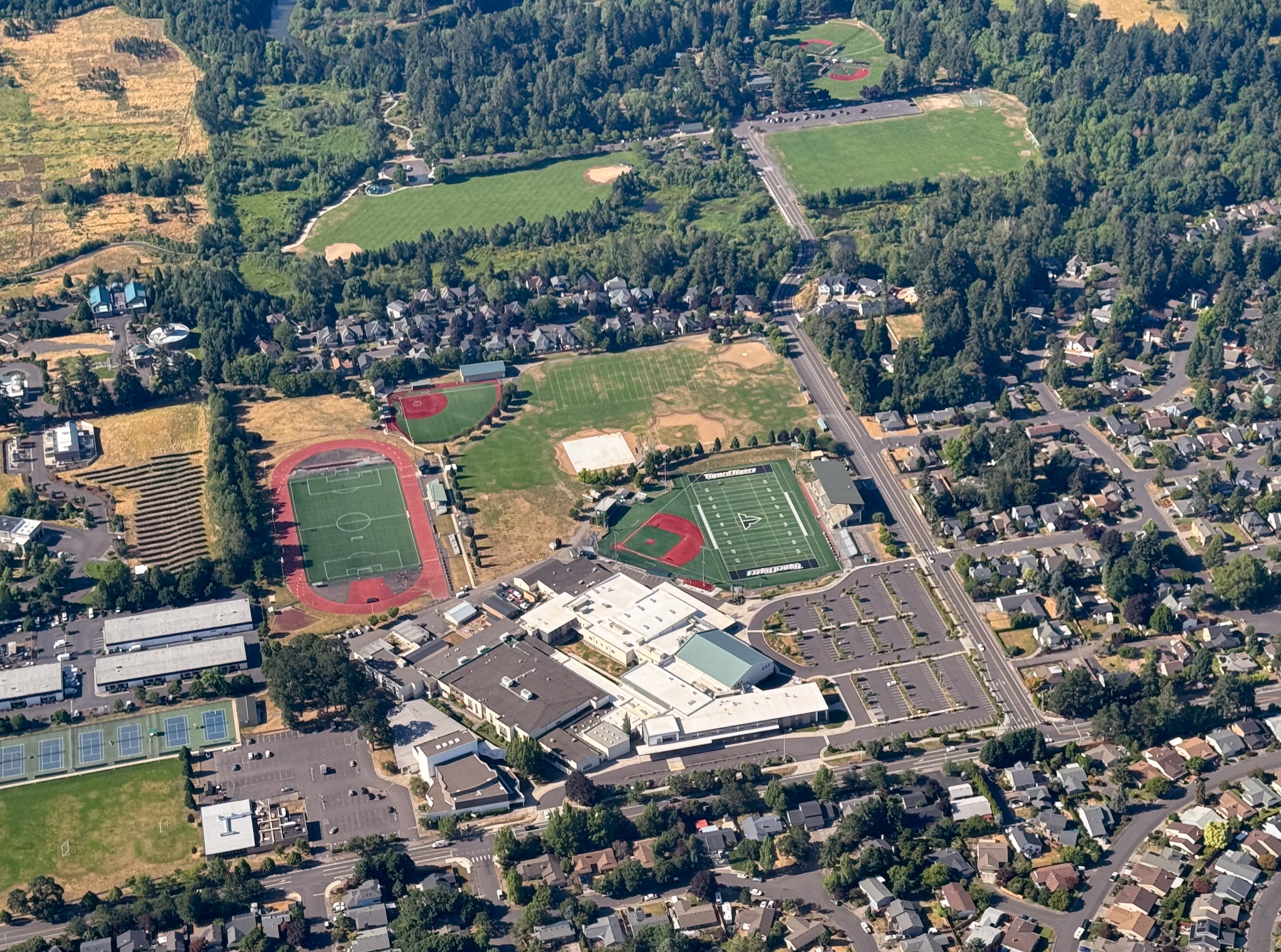 Aerial view of a suburban area with a large school complex featuring multiple sports fields, including a football field, baseball diamonds, tennis courts, and a running track, surrounded by residential neighborhoods and wooded areas.