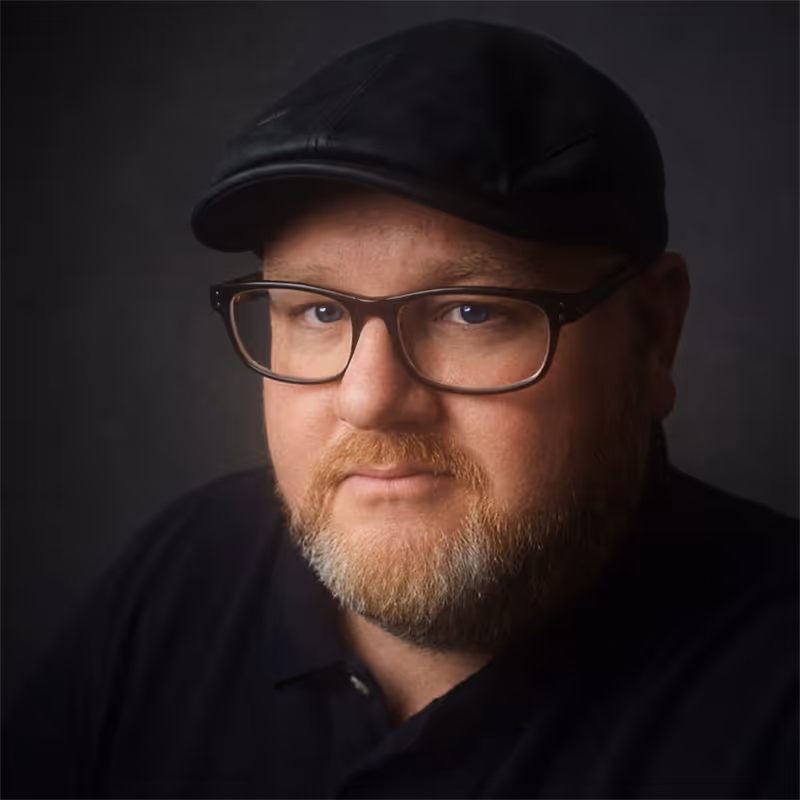 Man with light beard and glasses wearing a black cap and dark shirt against a dark background.