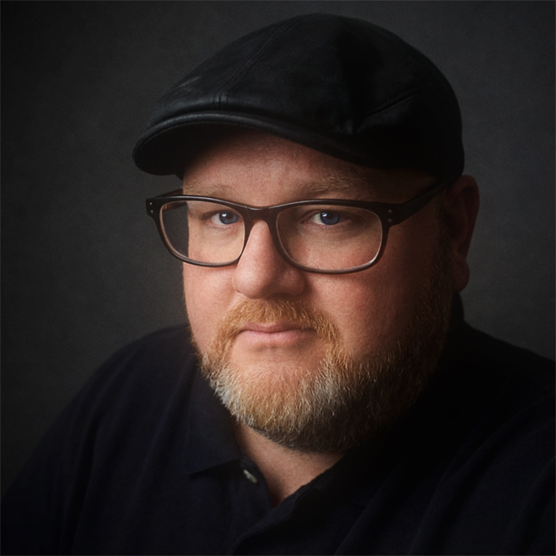 Man with light beard and glasses wearing a black cap and dark shirt against a dark background.
