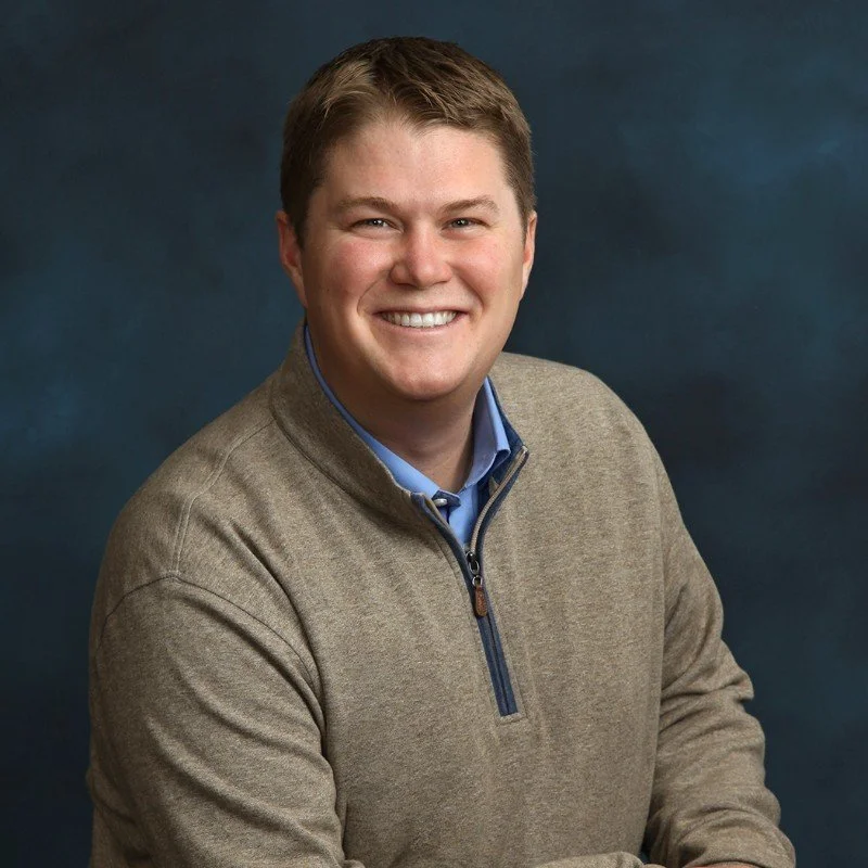 Smiling man named Logan Haugen with short brown hair wearing a beige quarter-zip sweater over a blue collared shirt against a dark blue background.