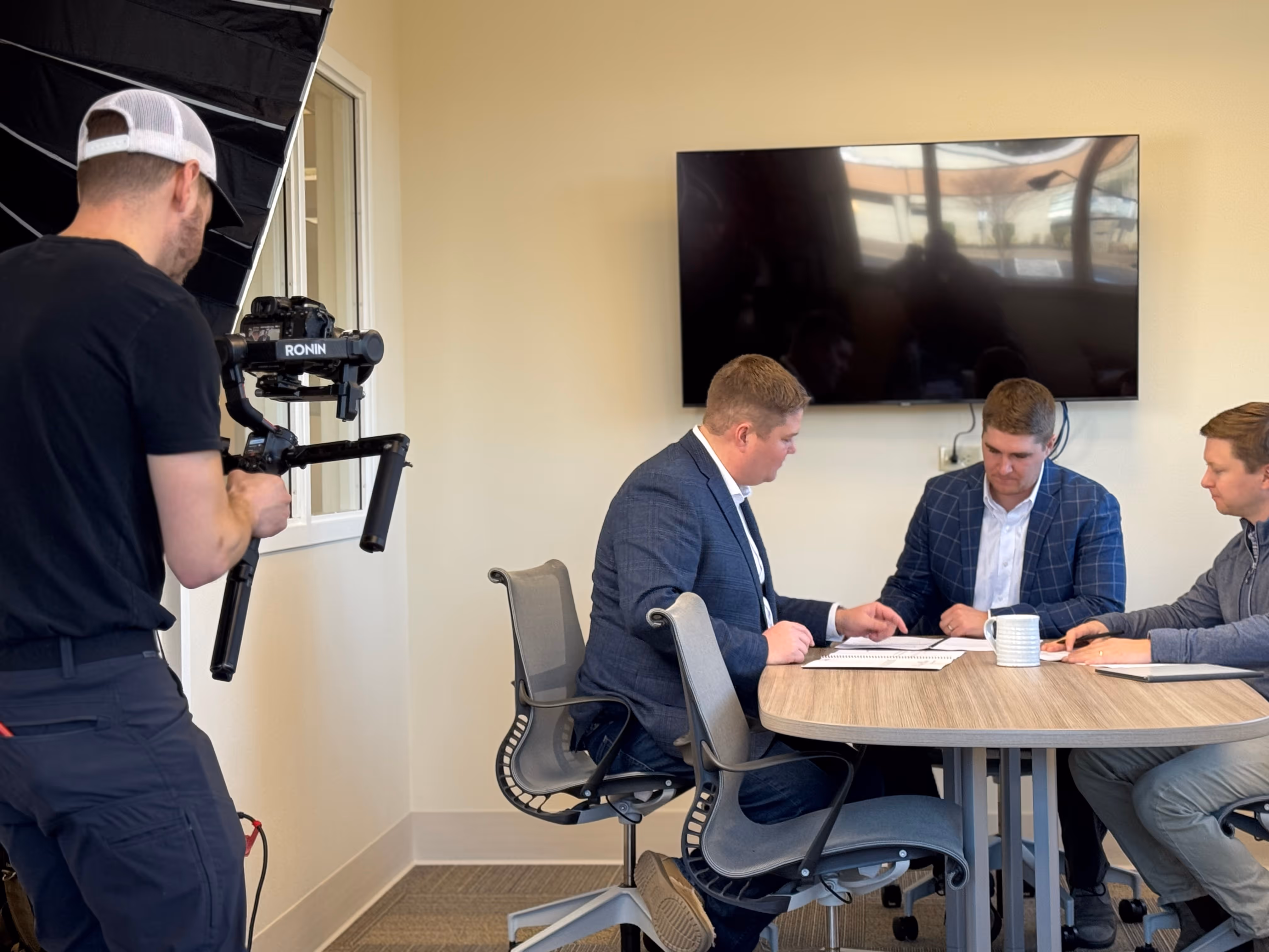Videographer recording three men in business attire discussing documents at a round conference table in an office with a large TV screen on the wall.