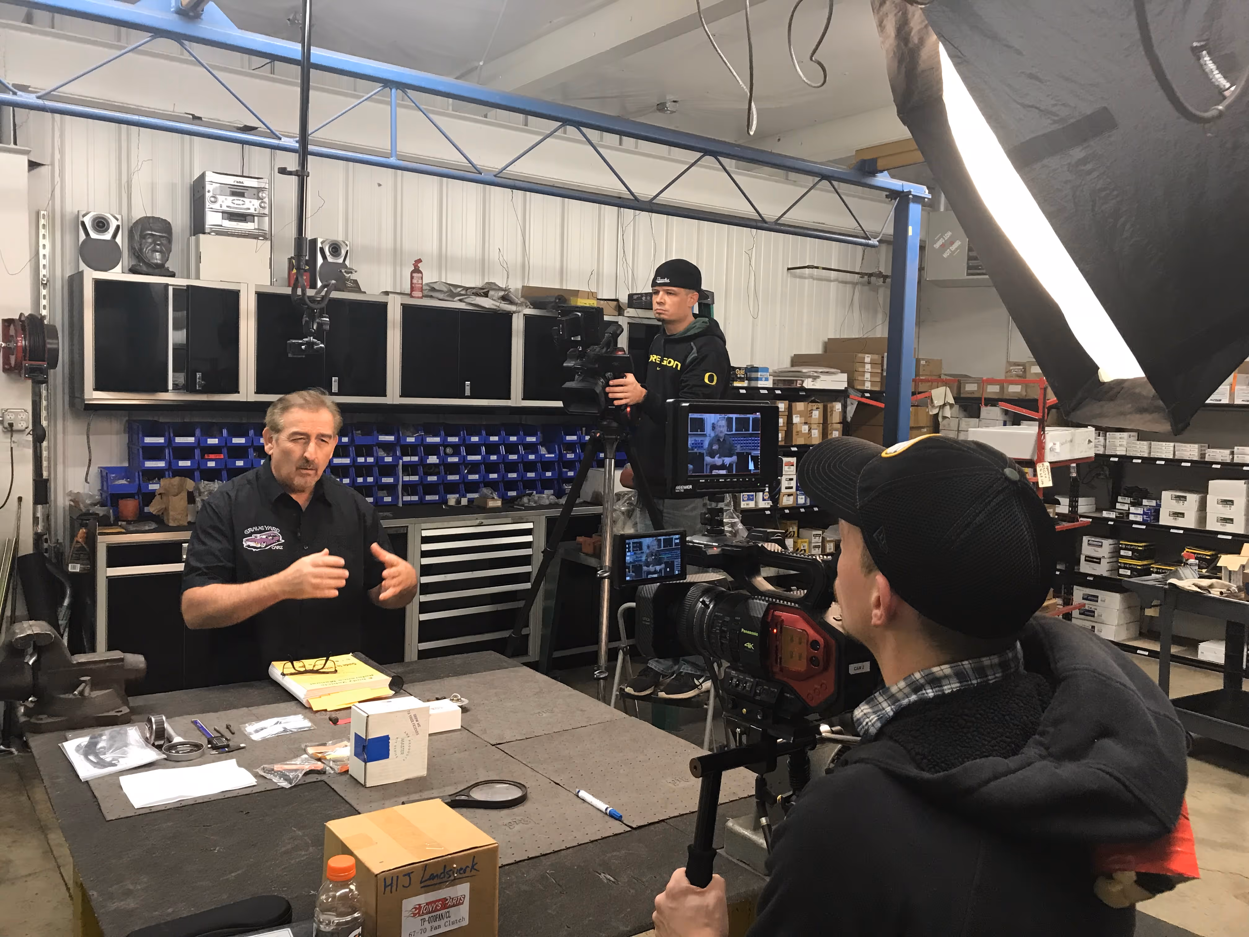 Man explaining at a workshop table with tools, filmed by two cameramen using professional video cameras and lighting.
