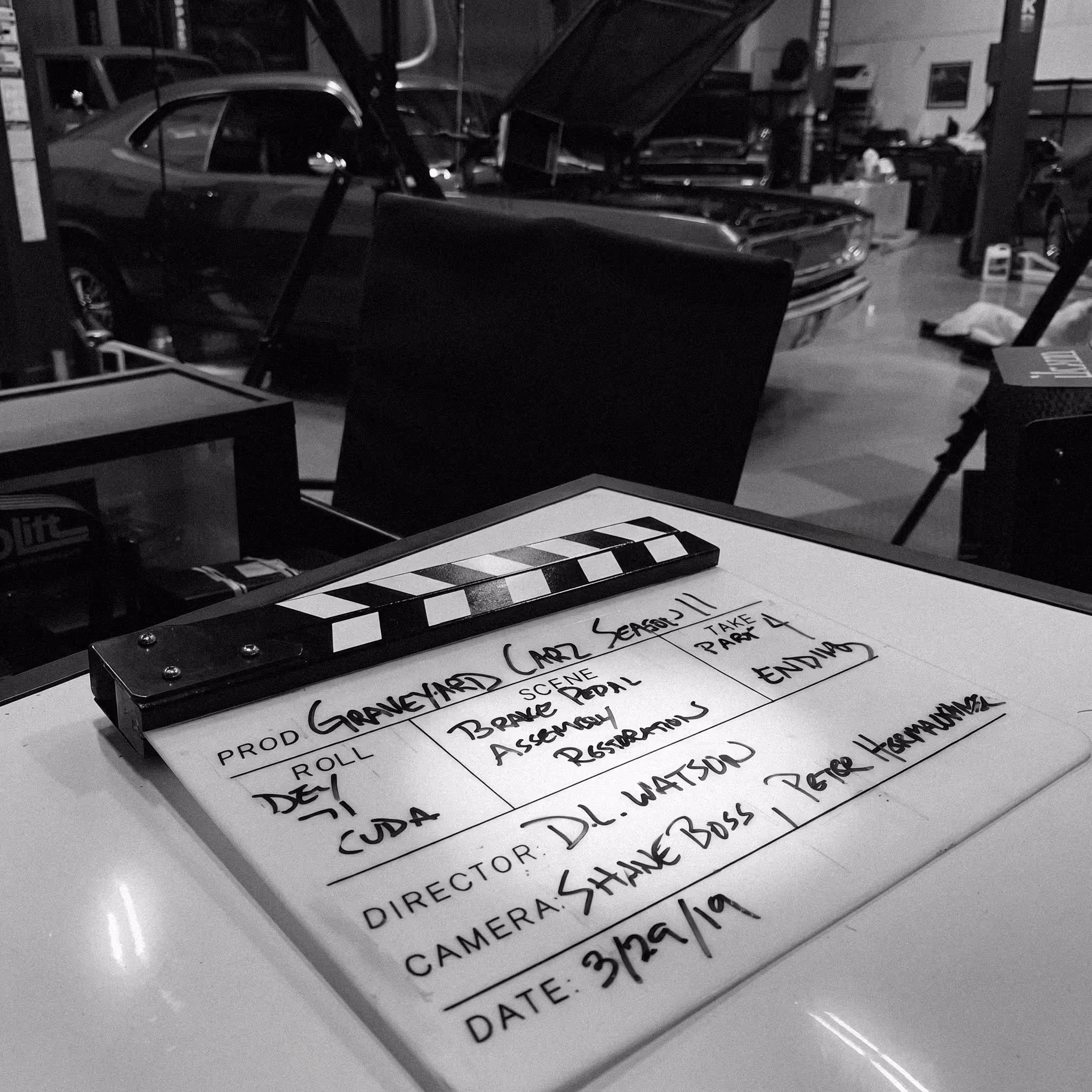 Black and white image of a film clapperboard with the directors name of "D.L. Watson" on a white surface in a garage, with classic cars and equipment in the background.