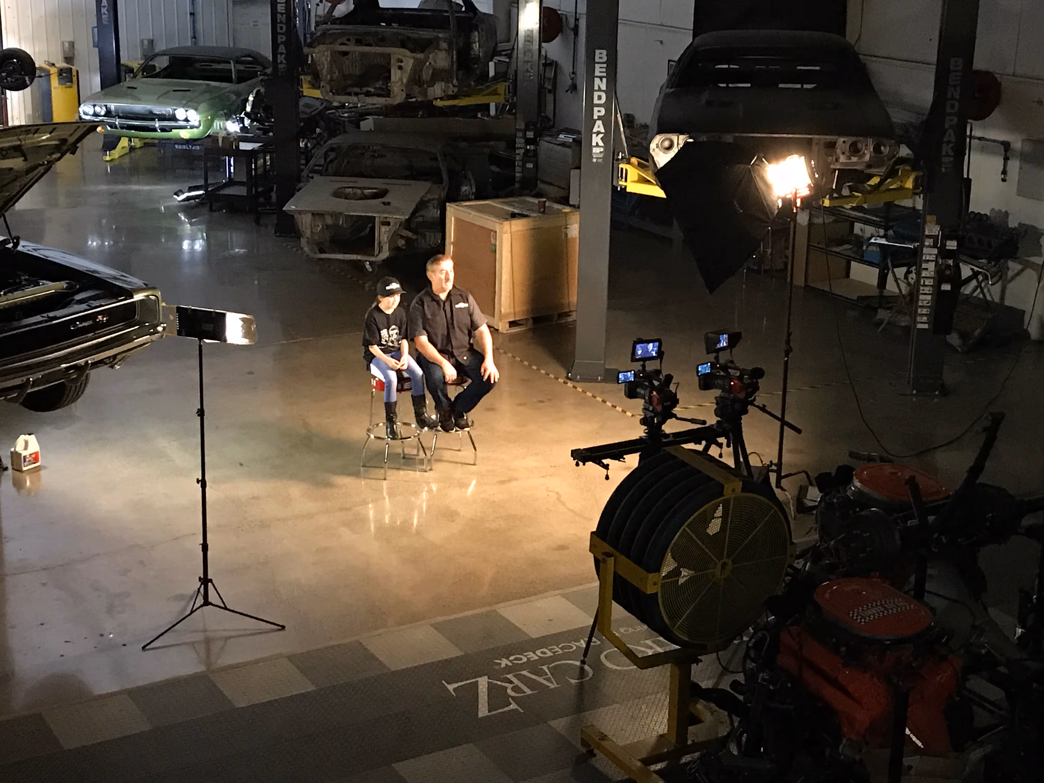 Two people sitting on stools under studio lights inside a large automotive workshop with cars and engines in the background, on set of Graveyard Carz, produced in Springfield, Oregon.