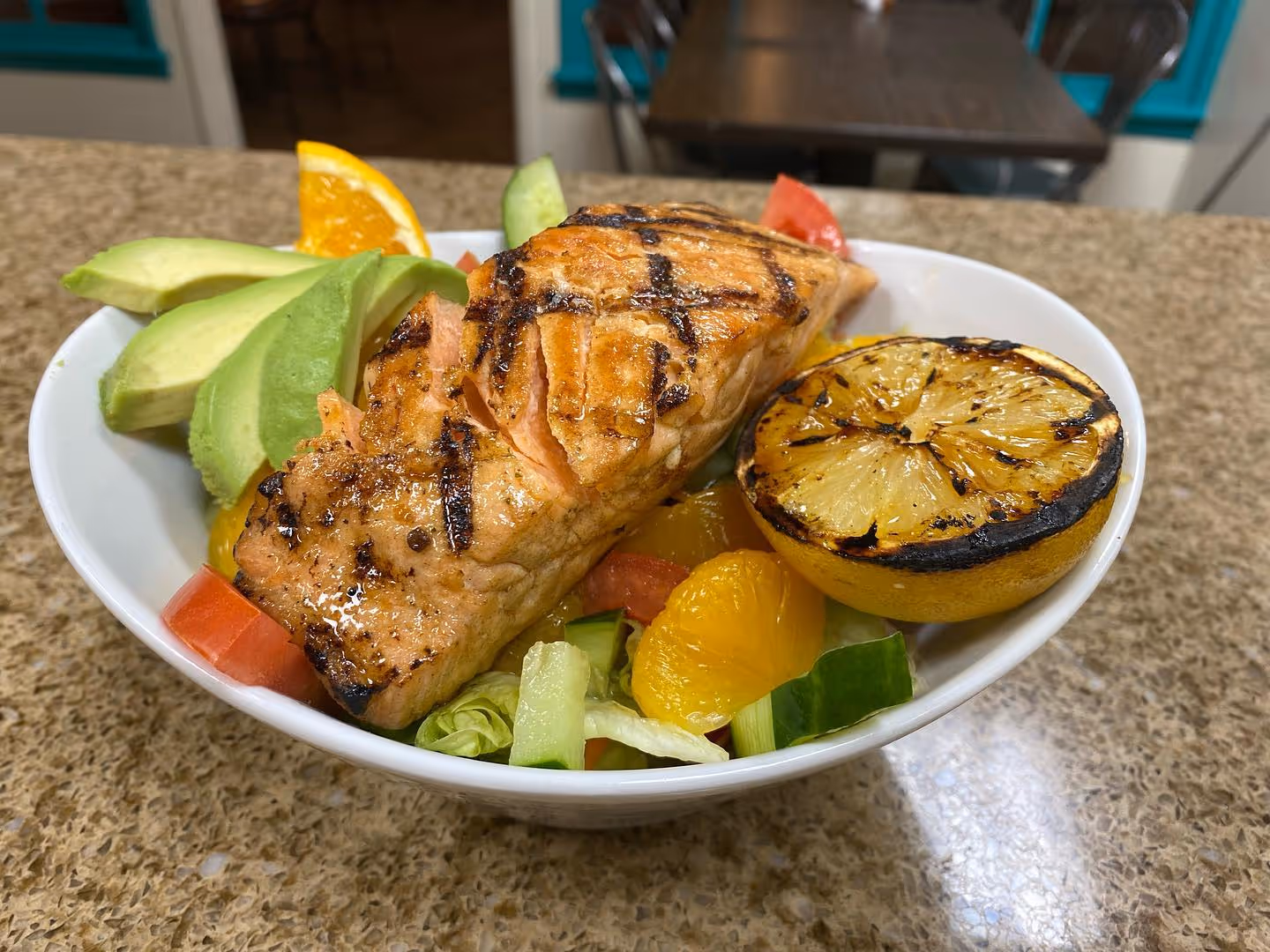 Grilled salmon fillet with slices of avocado, grilled lemon half, orange wedge, and mixed salad in a white bowl on a countertop.