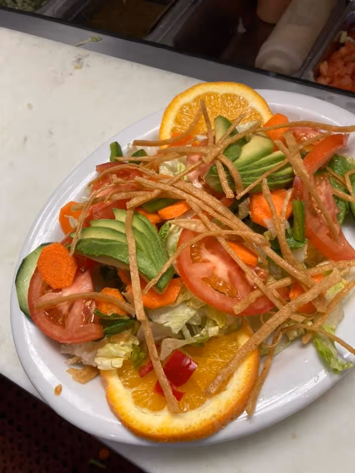 Fresh vegetable salad with sliced avocado, tomatoes, carrots, cucumber, lettuce, crispy tortilla strips, and orange slices on a white plate.