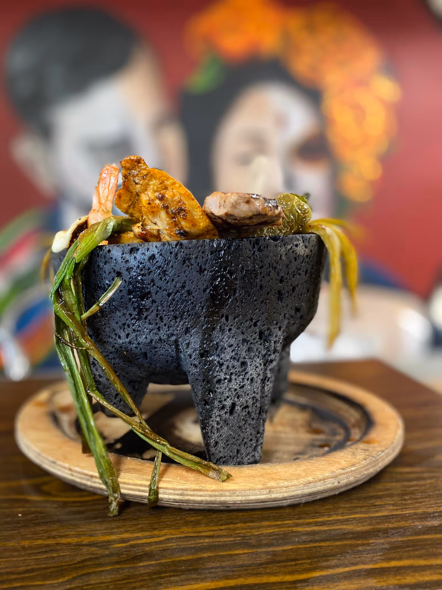 A stone molcajete bowl filled with grilled shrimp, chicken, steak, and roasted vegetables served on a wooden plate.