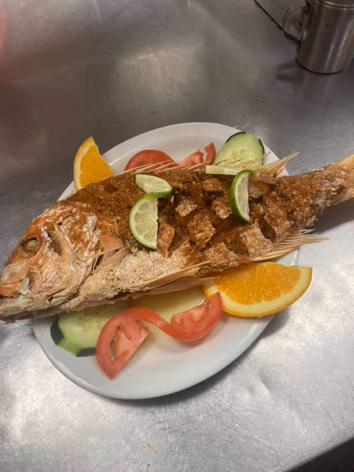 Whole fried fish garnished with lime slices, served on a white plate with slices of tomato, cucumber, and orange.